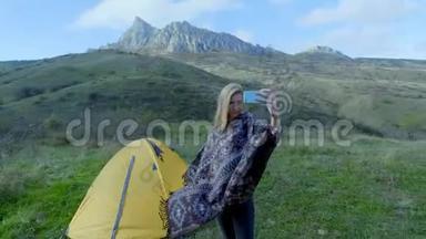 迷人的女孩徒步旅行使自拍在智能手机的背景山区。 一个女孩站在帐篷附近露营